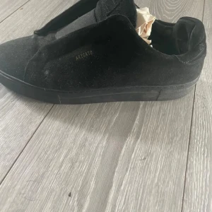 Axel Arigato skor - Stilrena svarta sneakers från Axel Arigato i mocka med diskret logga på sidan. Skorna har en platt sula och minimalistisk design som passar perfekt till streetwear. Klassisk rund tå och helsvart look för en clean vibe.