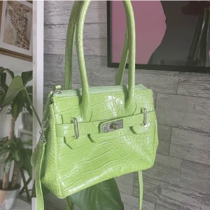 Grön croco handväska med axelrem - Säljer en limegrön handväska i croco-mönstrat syntetmaterial. Väskan har både handtag och avtagbar axelrem, silverfärgade metalldetaljer och snyggt spänne framtill. Perfekt för dig som vill sticka ut med en färgglad accessoar💚