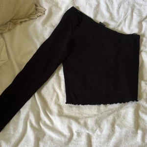 Svart oneshoulder topp med lång ärm - Säljer en svart oneshoulder topp med lång ärm och croppad passform. Toppen har en stilren look med vågig kant nertill och är perfekt för dig som gillar trendiga detaljer. Enkel att matcha med både jeans och kjol från ginatricot 