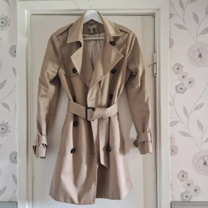 Beige trenchcoat med bälte - Stilren beige trenchcoat med klassisk dubbelknäppning, bred krage och bälte i midjan. Jackan har långa ärmar med spännen vid handlederna och svarta knappar framtill. Perfekt för dig som vill ha en tidlös och snygg look. Sitter bra även på dig som vanligtvis är strl 36. 