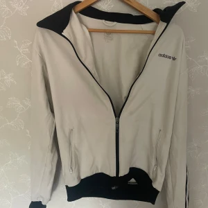 Vit Adidas vindjacka med svarta detaljer - Vit Adidas vindjacka med svarta detaljer och klassiska tre ränder längs ärmarna. Jackan har dragkedja framtill, två fickor med dragkedja och svart krage. Adidas-logga broderad på bröstet. Perfekt för en sportig och clean look.