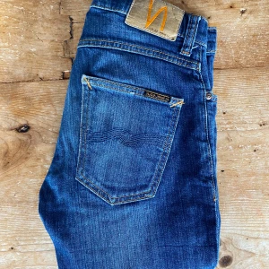 Blå Nudie Jeans, W27 L30 - Snygga blå jeans från Nudie Jeans med klassisk femficksdesign och orangea sömmar. Jeansen slim passform, tillverkade i slitstark bomullsdenim. Perfekta för dig som gillar en tidlös och stilren look. MODELL: Grim Tim                                           W:27  L:30