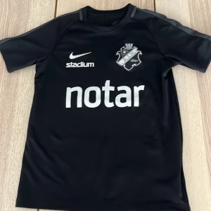 Svart AIK fotbollströja Nike M - AIK fotbollströja i svart från Nike med Dri-Fit-material. Tröjan har korta ärmar, klubbmärke på bröstet och tryck från Notar, Stadium och Svea Ekonomi. Perfekt för dig som vill visa ditt AIK-stöd på planen eller läktaren.