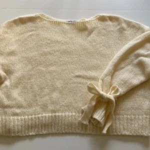 Krämvit stickad tröja med knytbar rosett - Superfin krämvit stickad tröja från Meet Me There. Tröjan har långa ärmar med snygg knytbar rosett vid ärmslutet och ribbade muddar. Aldrig använd 