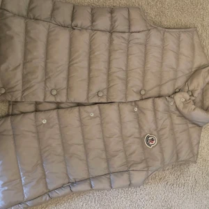 Beige dunväst från Moncler - Snygg beige dunväst från Moncler med klassisk quiltad design och tryckknappar framtill. Västen har hög krage och ikonisk Moncler-logga på bröstet. Perfekt att slänga över en hoodie eller tröja för extra värme och stil.