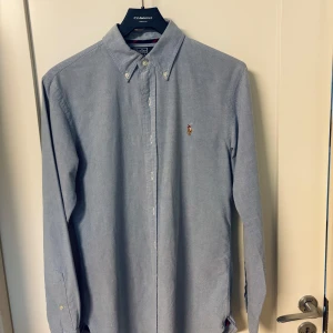 Blå oxfordskjorta - Ralph Lauren - Klassisk ljusblå skjorta från Polo Ralph Lauren i slim fit-modell, storlek medium. Skjortan har button down-krage, långa ärmar och broderad logga på bröstet.