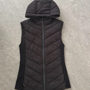 Svart dunväst med huva från Zara - Säljer en svart dunväst från Zara med quiltad design och dragkedja framtill. Västen har en stor, mysig huva och smala svarta sidopaneler som ger en snygg siluett. Perfekt att slänga över en hoodie eller tröja när det är kyligt ute.