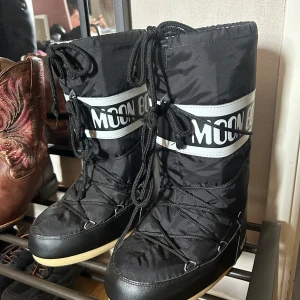 Svarta Moon Boots storlek 36/38 - Svarta Moon Boots med vit logga och snörning framtill. Yttermaterial i syntet och insida i mjukt, ljust foder. Perfekta för kalla vinterdagar och har en klassisk, chunky siluett med platt sula.