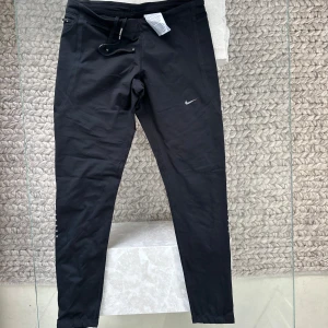 Svarta Nike träningsbyxor med dragkedja - Svarta träningsbyxor från Nike med smal passform och snyggt reflekterande dragkedja vid bensluten. Byxorna har snörning i midjan och är tillverkade i ett stretchigt funktionsmaterial som andas. Perfekta för löpning eller gymmet. Storlek M - passar som Storlek S.