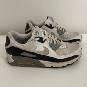 Nike Air Max 90 vita och marinblå sneakers - Nike Air Max 90 sneakers i vitt och marinblått med grå detaljer. Klassisk siluett med mesh och skinnpaneler, synlig Air Max-dämpning i sulan och snörning framtill. Perfekta för dig som gillar retro vibes och streetstyle.