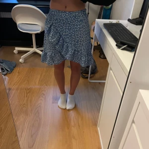 Blå blommig volangkjol - Supersöt blå kortkjol med vitt blommönster och volang. Kjolen har en smickrande passform och stängs med dragkedja i sidan.