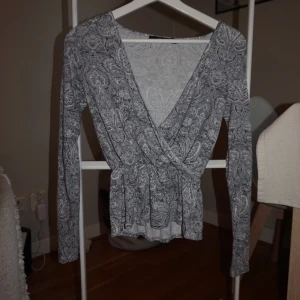 Grå omlottopp med paisleymönster BikBok - Supersnygg grå omlottopp från BikBok i storlek XS. Toppen har ett detaljerat paisleymönster, lång ärm och markerad midja. Djup v-ringning framtill ger en trendig look. Perfekt att styla med jeans eller kjol för en cool vibe.