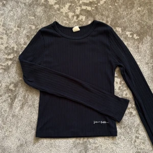 Svart ribbad långärmad tröja Urban Outfitters - Svart ribbad långärmad tröja från Urban Outfitters med broderad logga nertill. Tröjan har en rund halsringning och är i mjukt, stretchigt material som sitter snyggt på kroppen. Hör av dig vid frågor🩷