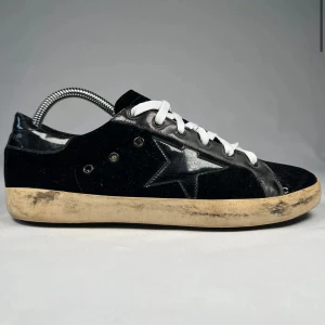 Svarta golden goose - Svarta sneakers med vit sula och vita snören. Skorna har en tydlig svart stjärna på sidan och detaljer i både mocka och skinn. Sulan är beige och har en sliten look som ger en cool vintage vibe. Perfekta för dig som gillar streetstyle.