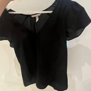 Svart blus med v-ringning - Enkel och stilren svart blus med v-ringad hals och korta, lite fladdriga ärmar. Sval och luftig känsla, perfekt till jeans eller kjol. Tidlös design som funkar till många olika looks.