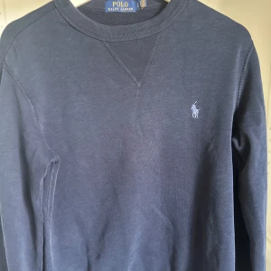 Mörkblå sweatshirt från Polo Ralph Lauren - En stilren mörkblå sweatshirt från Polo Ralph Lauren med klassisk rund halsringning och broderad logga på bröstet. Tröjan har långa ärmar och är tillverkad i mjuk bomull för en skön känsla. Perfekt för en avslappnad och snygg look. Perfekt till dig som vill börja resella eller ha märkeskläder till feta priser!
