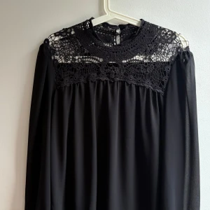 Svart bluss med spetsdetaljer - Svart långärmad bluss med vacker spets upptill. Blusen har knappar i nacken och ett flowy fall. Perfekt för dig som gillar romantiska detaljer och en stilren look.