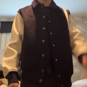 Axel Arigato bomberjacka  - Snygg Axel Arigato bomberjacka i svart och beige med broderad text på ryggen och knappstängning framtill. Jackan har kontrasterande ärmar och klassisk college-look. Perfekt för dig som vill sticka ut med en cool streetstyle. Nypris över 5000kr