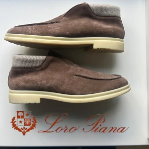 Loro Piana Open Walk - Bruna mockaskor från Loro Piana med beige ribbstickad kant och vit sula. Insidan är fodrad med mjukt material för extra komfort. Skorna har en stilren och minimalistisk design med rund tå och platt sula, perfekta för höst och vinter.