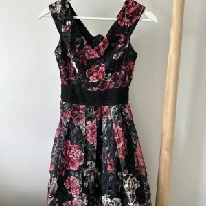 Blommig svart klänning med markerad midja - Supersnygg svart klänning med stora blommor i rosa, rött och beige. Klänningen har breda axelband, markerad midja med svart bälte och ett vackert veckat kjolparti. 