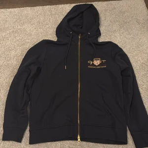 Marinblå hoodie från GANT med dragkedja - Snygg marinblå hoodie från GANT med broderad logga och text på bröstet. Tröjan har huva med snörning, hel dragkedja i guld och långa ärmar. Perfekt för en sportig och avslappnad look. Använd mycket fåtal gånger. Nypris 1700kr, nu 200kr. 