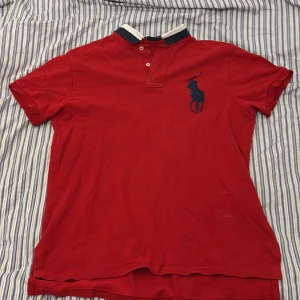 Röd Polo Ralph Lauren t-shirt M - Röd t-shirt från Polo Ralph Lauren med klassisk pikékrage, knappar och stor marinblå logga på bröstet. Custom fit-modell med korta ärmar och vit/marinblå detalj på kragen. Perfekt för en clean och sportig look. Lite märken vid magen som visas på bilden