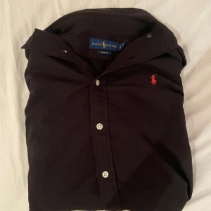 Svart skjorta från Ralph Lauren - Svart skjorta från Ralph Lauren i slim fit-modell. Klassisk design med knappar framtill och röd broderad logga på bröstet. Skjortan har lång ärm och är tillverkad i bomull, vilket gör den både bekväm och stilren.