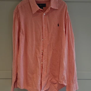 Rutig orange skjorta från Ralph Lauren - Snygg orange och vit rutig skjorta från Ralph Lauren med klassisk krage och knäppning framtill. Skjortan har lång ärm och ikonisk broderad logga på bröstet. Perfekt för dig som gillar stilren och tidlös design.