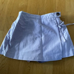 Ljuslila rutiga shorts från Zara - Säljer ett par ljuslila rutiga skorts från Zara i bomull. Shortsen har omlott-look framtill med knytband och knapp i midjan samt resår baktill för extra komfort. Perfekta för varma sommardagar och har en söt, avslappnad stil.