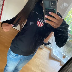 Svart sweatshirt från GANT med logga - Svart sweatshirt från GANT med broderad logga och sköld med stjärnor och ränder på bröstet. Klassisk rund halsringning och långa ärmar. Mjuk och skön bomullsmix, perfekt för en avslappnad stil.