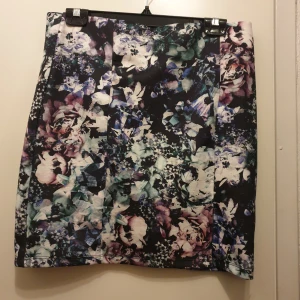 Blommig kjol från Gina Tricot - Supersnygg kjol från Gina Tricot med blommigt mönster i svart, lila, blått och vitt. Kjolen har en liten slits på sidan och är gjord i ett mjukt, stretchigt polyestertyg. Aldrig använd. Storlek L 