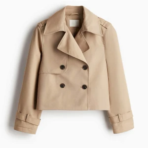 Kort beige trenchcoat  - Snygg croppad trenchcoat i beige med dubbelknäppning och stora slag. Jackan har bältdetaljer vid ärmsluten och klassisk krage.                  Lägger ut för att se intresse då den inte kommit till användning, slutsåld på hemsidan