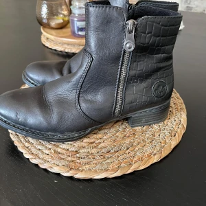 Svarta boots från Rieker, storlek 38 - Säljer ett par svarta boots från Rieker i storlek 38. Skorna har en rund tå, platt sula och dragkedja på sidan. Ovandelen är i skinn med en snygg krokodilpräglad detalj på skaftet. Perfekt för dig som gillar stilrena och coola detaljer.