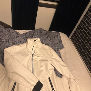 Vit vindjacka från Race Marine - Snygg vit vindjacka från Race Marine i storlek M. Jackan har svart dragkedja och svarta detaljer vid fickorna och ärmsluten. Hög krage och stilren design, perfekt för dig som gillar sportig och clean look. Materialet är lätt och syntetiskt.