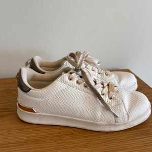 Vita sneakers från Elle Sport, strl 39 - Snygga vita sneakers från Elle Sport med textur i reptilmönster och detaljer i guld. Skorna har snörning, rund tå och en platt sula. Bakkappan är grå med guldfärgade nitar och en metallicrand runt hälen. Perfekt för dig som gillar stilrena och trendiga sneakers.
