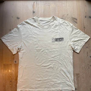 Vit t-shirt med tryck från Jack & Jones - Snygg vit beige från The Gallery (Jack & Jones) med coolt tryck fram och bak. Framsidan har svart text och grafisk tryck, baksidan har färgglada druvor i gult och rött samt stor text. Skön passform och tillverkad i bomull. Skriv vid intresse!
