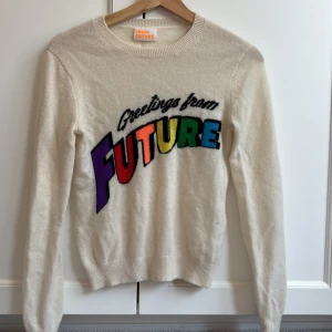 Vit kashmirtröja från From Future - Säljer en mjuk och lyxig vit kashmirtröja från From Future med färgglad text 'Greetings from FUTURE' i regnbågens färger på bröstet. Rund hals och långa ärmar, perfekt för dig som gillar statement-plagg och vill sticka ut med coola färger.
