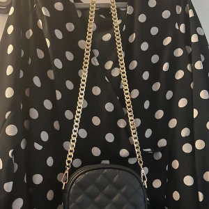 Svart quiltad axelväska med guldkedja - Snygg, svart liten axelväska  i quiltad skinnimitation. Liknar Pom Pom London.  Väskan har en oval form, guldfärgad kedja som axelrem och dragkedja upptill. Och en liten avtagbar Pom Pom boll som dekoration. Innerficka. Perfekt storlek för mobilen och det viktigaste. Liten metalllogga framtill. Bra skick, knappt använd! 