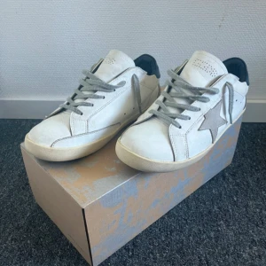 Golden Goose Super-Star sneakers - Golden Goose Super-Star sneakers i vit skinn med grå mockadetaljer och stjärna på sidan. Skorna har grå snörning, rund tå och beige sula. Klassisk low-top modell med perforering på tungan och svart häl. Perfekt för dig som gillar italiensk streetstyle.