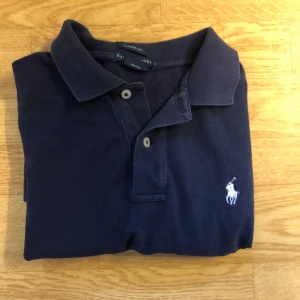 Marinblå polo t-shirt från Ralph Lauren - Snygg marinblå polo t-shirt från Ralph Lauren i klassisk passform. T-shirten har korta ärmar, krage och två knappar framtill. Ikoniska vita Polo-loggan broderad på bröstet. Perfekt för en clean och stilren look. Storlek L passar M