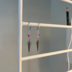 Silvriga örhängen med rosa pärla - Coola örhängen med silvriga konformade hängen och en liten rosa pärla ovanför. Perfekt för dig som gillar edgy detaljer och vill sticka ut lite extra. Snygga att matcha med både streetwear och mer stilrena outfits. I rostfritt stål och handgjort