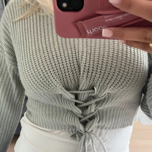 Ljusgrå croppad stickad tröja med snörning - Superfin ljusgrå croppad stickad tröja med breda ärmar och snygg snörning framtill. Tröjan har ett grovt ribbat mönster och sitter tajt i midjan. Perfekt att styla med jeans eller kjol för en trendig look.