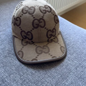 Gucci beige keps med GG-mönster - Säljer en snygg beige keps från Gucci med det klassiska GG-logomönstret i brunt över hela kepsen. Kepsen har böjd skärm, justerbar rem bak och är tillverkad i slitstark bomull. Perfekt för dig som vill ha en lyxig och trendig accessoar.