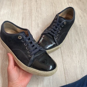 Svarta lanvin cap toe  - Snygga svarta lanvin sneakers med cool lackad tå och ovandel i mocka. Skorna har snörning och en beige platt sula som ger en klassisk vibe. Perfekta för dig som gillar stilrena detaljer och vill sticka ut med en unik look. De är i ganska bra skick,skriv till mig vid minsta fundering.Ingen og medföljer tyvärr. Några säger att de ser marinblå ut men jag tycker att de ser i mestadels svarta ut.Skriv till mig när ni ska köpa.🤔 😃💸 Mvh Emil