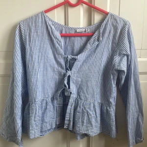 Blåvit randig blus från Name It - Blus i blått och vitt med smala ränder från Name It. Modellen har långa ärmar, volang nedtill och snygg snörning framtill. Luftigt material som känns skönt mot huden, perfekt för vår och sommar. 