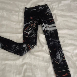 Svarta leggings från Stronger med mönster - Snygga svarta leggings från Stronger med coolt abstrakt mönster i orange, vitt och grått. De har två vita ränder på ena benet och elastisk midja med logga. Perfekta för träning eller chill. Materialet är stretchigt och känns skönt mot huden.