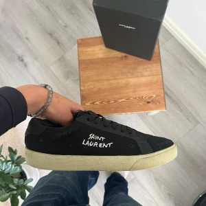 Svarta Saint Laurent sneakers strl 42 - TAR GÄRNA MOT BYTEN!                                    PRIS KAN FÖRHANDLAS🙏                                                                                        Svarta sneakers från Saint Laurent med vit logotyp på sidan och beige platt sula. Snygg, minimalistisk design med rund tå och snörning. Tillverkade i canvas och skinn för en cool och stilren look. Perfekt för dig som gillar exklusivt. 
