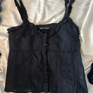 Svart blommig linnetopp från Zara - Supersnygg svart linnetopp från Zara med blommigt broderi och volangdetaljer längs axelbanden och knappraden. Toppen har smala axelband och en v-ringad hals. Perfekt att styla med jeans för en chill look.