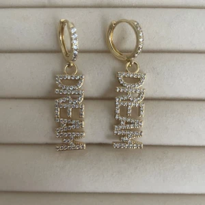 Glittriga örhängen med DREAM-text - Snygga örhängen i guldton med hängande DREAM-text, täckta av små glittrande stenar. Runda hoops med stenar som ger extra bling och cool vibe. Perfekt för dig som vill sticka ut och sprida good vibes med ditt smycke.