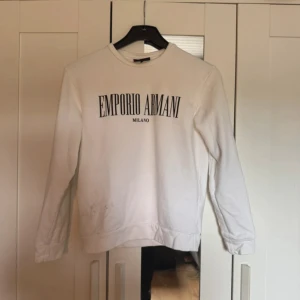 Vit sweatshirt Emporio Armani - Snygg vit sweatshirt från Emporio Armani med svart logotyptryck på bröstet. Tröjan har rund hals och långa ärmar, perfekt för dig som gillar stilrena och klassiska märkesplagg. Mjuk bomullskvalitet som är skön att bära.